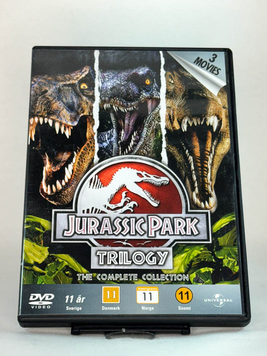 Forside av DVD-cover til Jurassic Park Trilogy – The Complete Collection – samleboks med alle tre filmene i Steven Spielbergs ikoniske dinosaurserie, brukt eventyrfilm på DVD hos Brukbar Film.