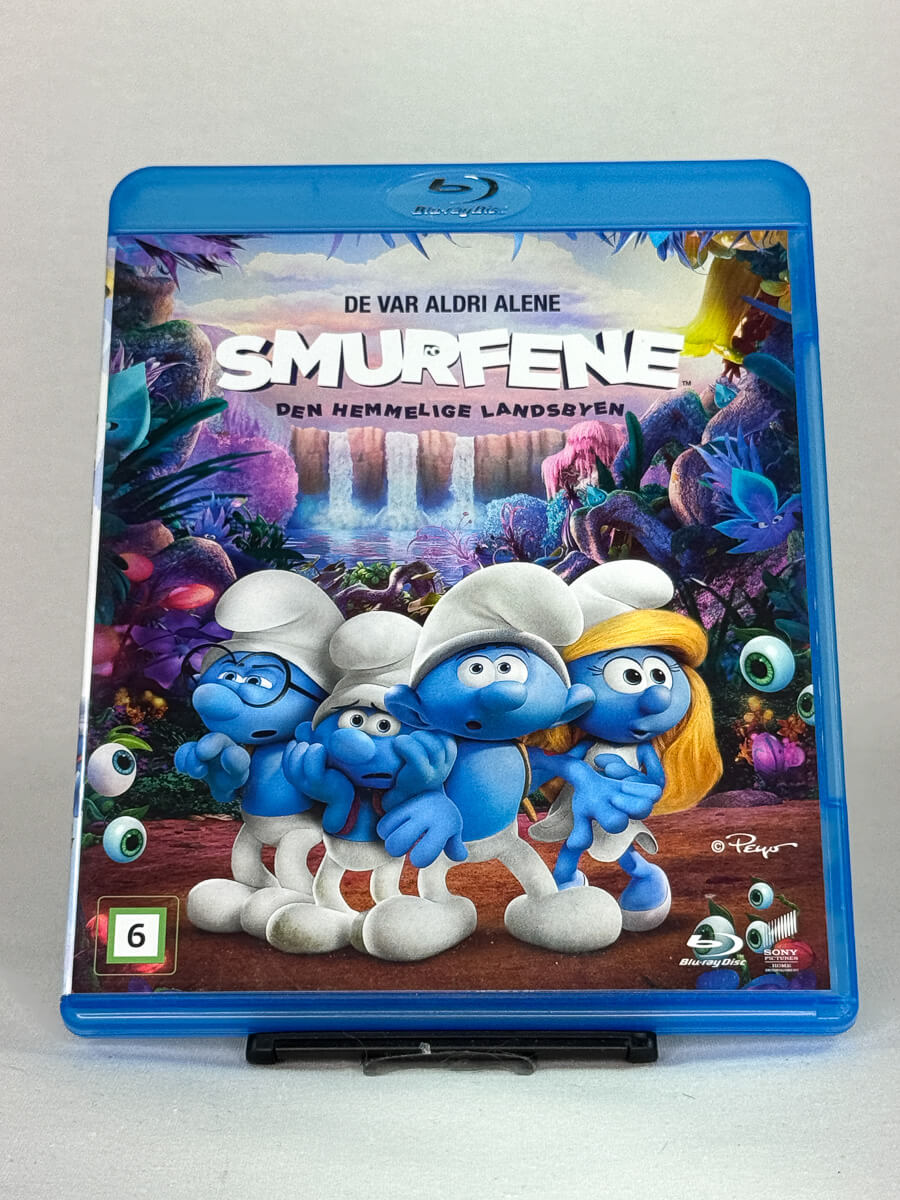 Forside av Blu-ray-cover til Smurfene – Den hemmelige landsbyen – fargerik animasjonsfilm der Smurfeline og vennene hennes drar på oppdagelsesferd, brukt Blu-ray hos Brukbar Film.