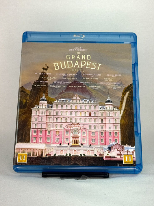 Forside av Blu-ray-cover til The Grand Budapest Hotel – Wes Andersons prisbelønte komedie med Ralph Fiennes og et stjernespekket ensemble, brukt Blu-ray hos Brukbar Film.